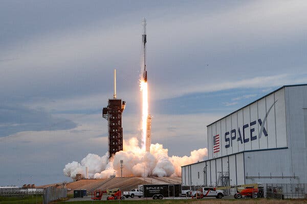 A rocket lifts off near a SpaceX facility with SpaceX branding on its side.