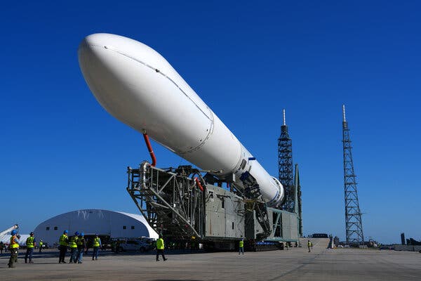 An enormous white rocket on a carrier makes its way very slowly to the launch stand.