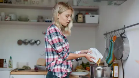 Getty Images A young woman with shoulder length blonde hair wearing a navy red and white check shirt pouring pasta into a silver pan on a stove in a basic looking kitchen