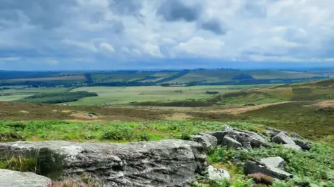 The Wildlife Trust/PA A picture showing the dramatic landscape of the Simonside Hills on the Rothbury Estate. In what appears to be a picture postcard of patchwork countryside, there are rocks at the front of the image with shrubbery. The sky is cloudy.