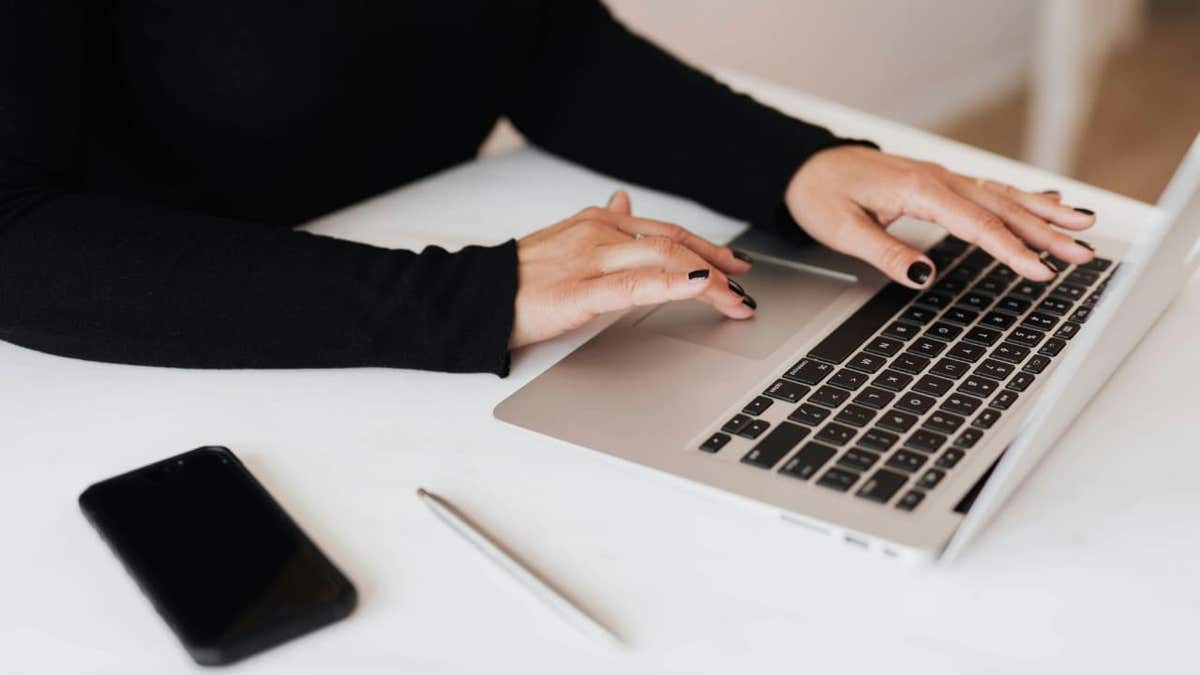 Person working on a MacBook at a white desk with a smartphone and pen nearby.