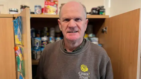 Chris Craddock/BBC Vinni Jones is wearing a greyish brown sweatshirt with a yellow rose logo and the words Grace Trust in white. He is standing in front of a brown cupboard which has piles of cans of food on the shelves. There are also colourful drawings on the cupboard door.