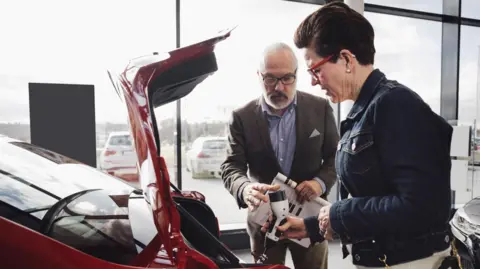 Getty Images Salesman in a car showroom shows plug connected to an electric vehicle to a female customer 